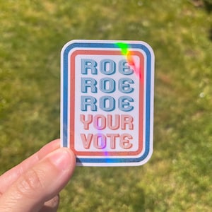 Puede incluir: Una pegatina con un borde rojo, blanco y azul y el texto "ROE ROE ROE YOUR VOTE" en una fuente retro.