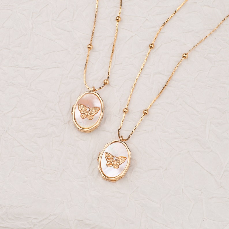 May include: Two gold-toned necklaces featuring oval-shaped lockets with a mother-of-pearl inlay. Each locket displays a textured gold butterfly emblem with a small clear stone at the center. The chains are delicate with evenly spaced gold beads.
