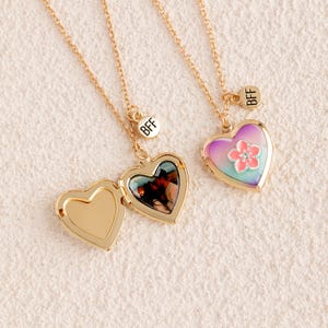 May include: Two gold-tone heart-shaped lockets on matching chains. One locket is open, revealing a photograph inside. The other locket is closed, featuring a gradient purple and blue enamel finish with a pink flower and a small clear stone. Both have a round charm with the text BFF.