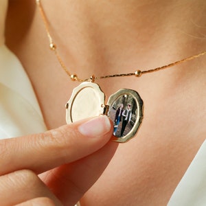 May include: A gold-toned oval locket necklace with a scalloped edge, suspended from a delicate chain with small bead accents. The locket is open, revealing a small, vertical photograph of two people standing together in an outdoor setting.
