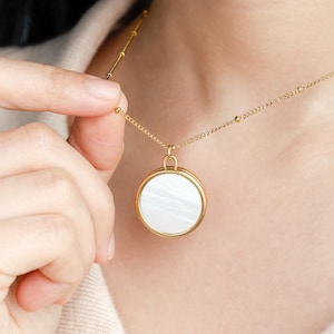 Pode incluir: Um colar de tom dourado com uma corrente delicada que apresenta pequenas contas espaçadas uniformemente e um pingente circular. O pingente tem uma moldura de metal dourado polido que envolve uma incrustação circular branca iridescente.