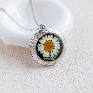 May include: A round silver-toned metal locket necklace featuring an embossed floral border pattern. The center of the locket displays a pressed white daisy flower with a yellow center set against a solid black background, suspended by a thin chain.
