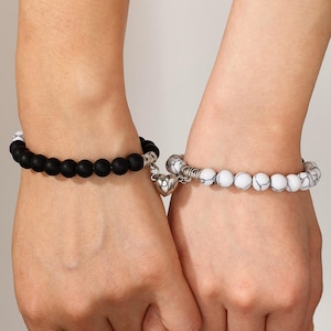 May include: Two beaded bracelets connected by a silver-toned magnetic heart charm engraved with the letters DK. One bracelet features matte black spherical beads, while the other consists of white beads with grey veining and silver-toned accents.