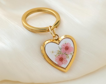 May include: A gold-coloured heart-shaped keyring with a clear centre displaying pink flowers. The keyring has a gold ring and a small clasp connecting the heart pendant. The background is a soft, white fabric.