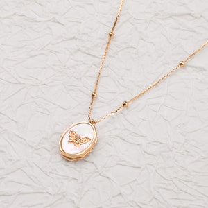 May include: A gold-toned necklace featuring a delicate chain with small spherical beads and an oval-shaped pendant. The pendant has a white, iridescent inlay with a central gold butterfly motif accented by a single small, clear gemstone.