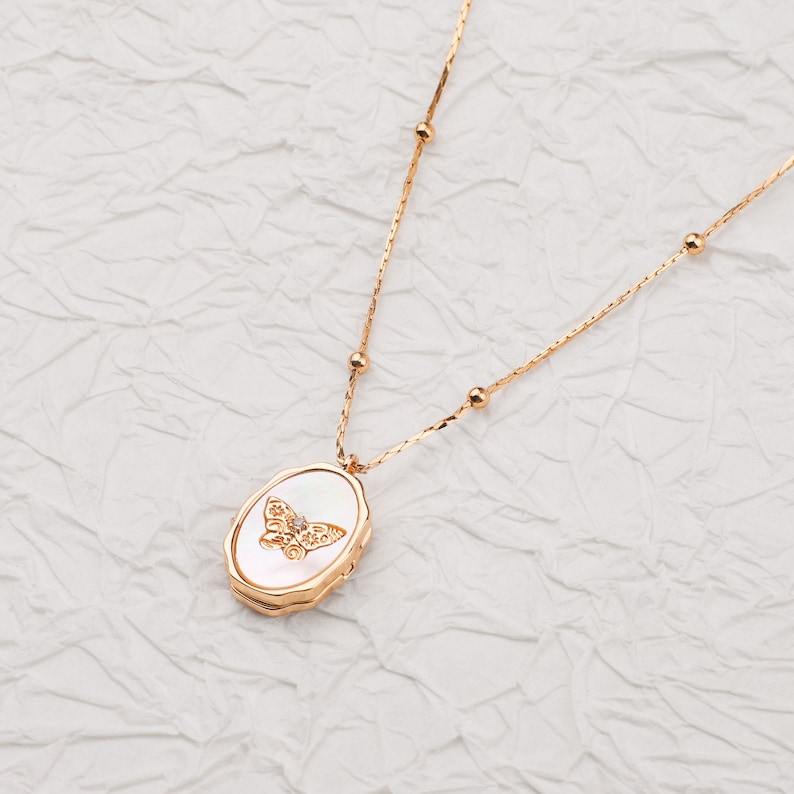 May include: A gold-toned necklace featuring a delicate chain with small spherical beads and an oval-shaped pendant. The pendant has a white, iridescent inlay with a central gold butterfly motif accented by a single small, clear gemstone.