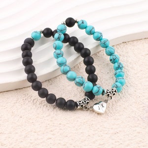 Puede incluir: Dos pulseras de cuentas conectadas por un dije en forma de corazón con las iniciales "J" y "K". Una pulsera está hecha de cuentas negras y la otra de cuentas turquesa.