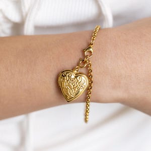 May include: A gold heart-shaped locket with a rose design hangs from a gold chain bracelet.
