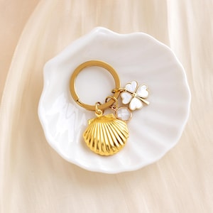 Op de afbeelding: Gouden sleutelhanger met een schelpbedel, een heldere kristallen bedel en een witte klavertje vier bedel.
