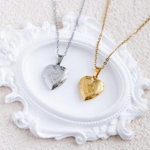 Puede incluir: Dos relicarios en forma de corazón sobre finas cadenas de metal. Un relicario es de tono plateado y el otro de tono dorado. Ambos relicarios presentan un patrón de borde decorativo grabado y un detalle central liso en forma de corazón.