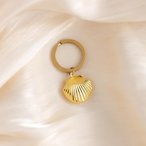 Puede incluir: Llavero de concha dorada con un anillo circular. El dije de concha tiene un diseño detallado y texturizado.