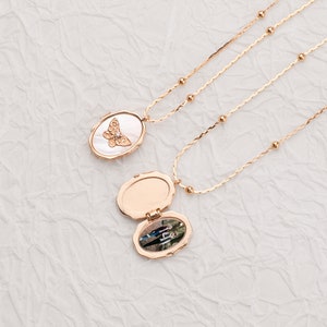 May include: Two rose gold-toned necklaces with delicate beaded chains. One oval locket is closed, featuring a white mother-of-pearl inlay with a gold butterfly motif. The second locket is open, revealing a small photograph of two people inside.