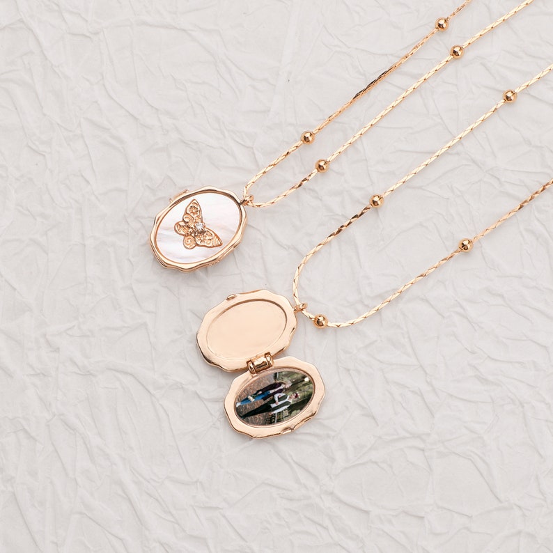 May include: Two rose gold-toned necklaces with delicate beaded chains. One oval locket is closed, featuring a white mother-of-pearl inlay with a gold butterfly motif. The second locket is open, revealing a small photograph of two people inside.