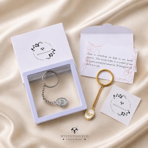 May include: Two keychains with oval pendants featuring a butterfly motif, one silver-toned and one gold-toned, each with a chain and ring. Includes a white gift box, a small envelope with a note, and two circular stickers with the text MYSTICVOGUECO.