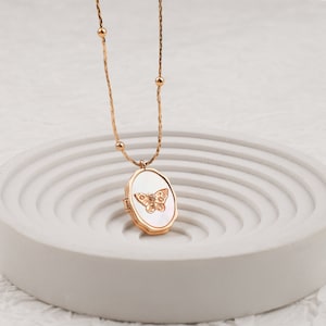 May include: A gold-toned necklace featuring a delicate chain with small spherical beads and an oval locket pendant. The locket has a white mother-of-pearl inlay centered with a gold-toned butterfly motif and a small clear stone at its center.