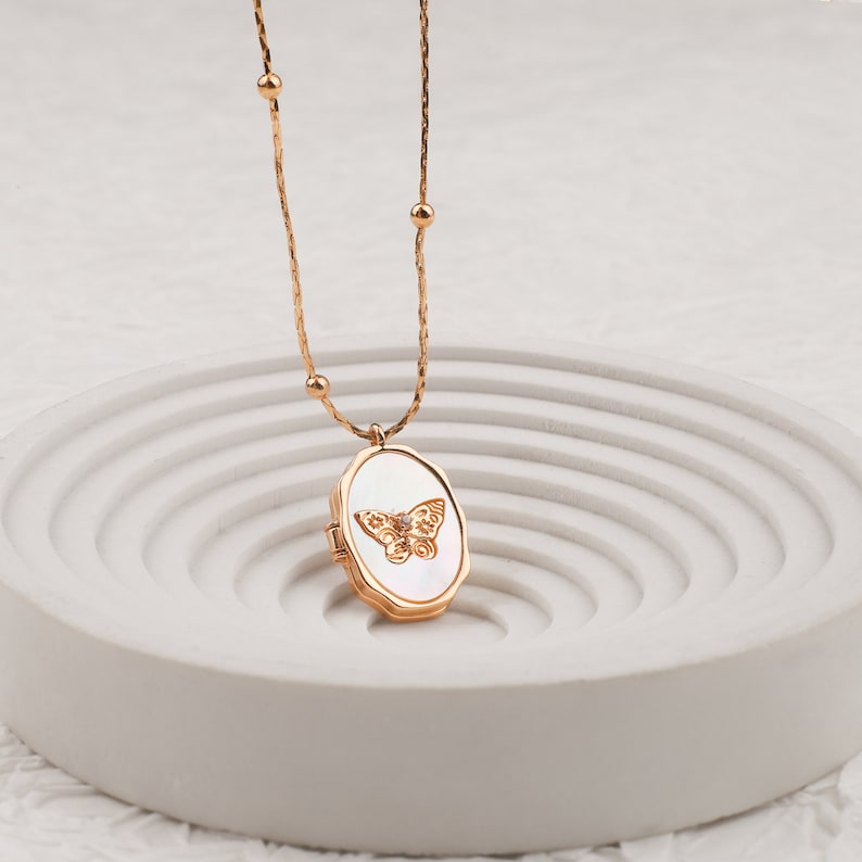 May include: A gold-toned necklace featuring a delicate chain with small spherical beads and an oval locket pendant. The locket has a white mother-of-pearl inlay centered with a gold-toned butterfly motif and a small clear stone at its center.