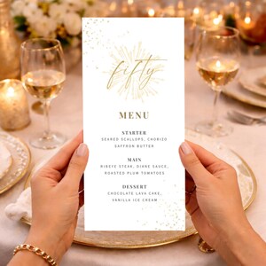 May include: A white menu card with gold text and accents, held by hands. The menu reads "fifty" at the top, followed by "MENU", and lists starter, main, and dessert courses. Wine glasses and a candle are in the background.