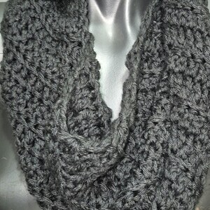 May include: A grey, hand-crocheted infinity scarf. The scarf is made of a thick, textured yarn, and is displayed on a mannequin. The scarf is a solid colour and appears to be warm and cosy. The style is suitable for autumn and winter.