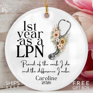 May include: A white ceramic ornament with a gold string for hanging. The ornament features the text "1st year as a LPN" and a stethoscope design with floral accents. The text "Proud of the work I do and the difference I make. Caroline 2026" is also included.