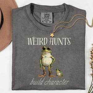 May include: Dark gray t-shirt with the words "WEIRD HUNTS" and "build character" in white script. The shirt features a frog wearing sunglasses and boots, with a smaller frog beside it. A gold star necklace is draped on the shirt.