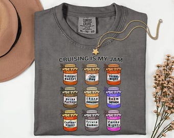 May include: Dark gray t-shirt with the text "CRUISING IS MY JAM" and illustrations of jars labeled with cruise activities like "Midnight Buffet" and "Casino Nights." A gold star pendant necklace is draped across the shirt.