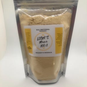 May include: A clear, resealable pouch filled with light brown powder. The pouch has a silver metallic top. A yellow label reads "Lion's Mane 100:1" and "Shaggy's Herbals". The label also states "All Natural Organic".