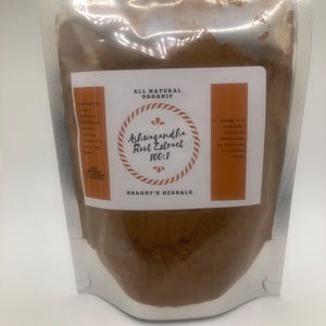 May include: A clear, resealable pouch filled with brown powder. The pouch has a white label with the text "ALL NATURAL ORGANIC" and "Ashwagandha Root Extract 100:1". The brand name "SHAGGY'S HERBALS" is also visible.
