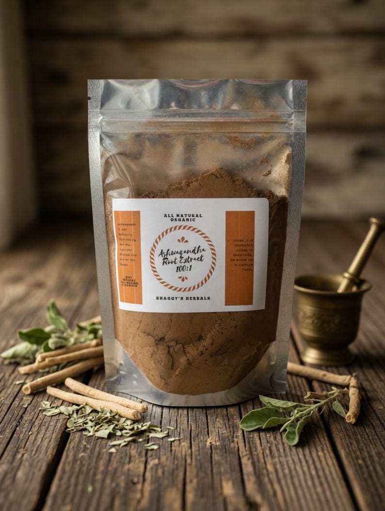 May include: A clear, resealable pouch filled with brown powder, labeled "Ashwagandha Root Extract 100:1" and "All Natural Organic." The pouch sits on a rustic wooden surface with sprigs of green leaves, stems, and a small brass mortar and pestle.