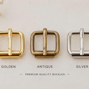 May include: Three square metal buckles arranged in a row, featuring a shiny golden finish, a matte antique brass finish, and a polished silver finish. The text below the buckles reads GOLDEN, ANTIQUE, SILVER, and PREMIUM QUALITY BUCKLES.