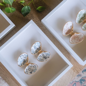 May include: Three white square frames with gold trim, each containing two decorative ceramic shells with floral patterns. The shells are in shades of green, pink, and blue.