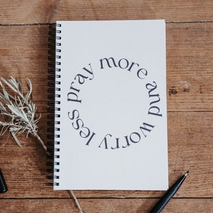 Könnte beinhalten: Ein weißes Notizbuch mit Spiralbindung und dem Text "worry less pray more" in einem Kreis auf dem Einband. Das Notizbuch liegt auf einer Holzoberfläche mit einem schwarzen Stift und einem Zweig getrockneter Kräuter.