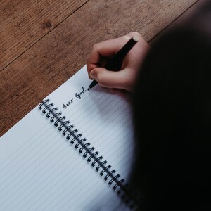 K&ouml;nnte beinhalten: Eine Person schreibt "Lieber Gott" in ein liniertes Notizbuch mit einem schwarzen Stift.