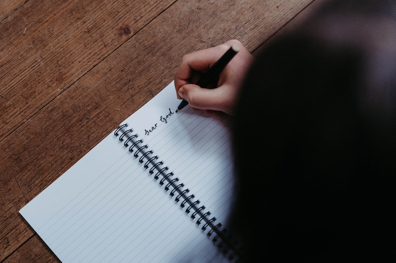 K&ouml;nnte beinhalten: Eine Person schreibt "Lieber Gott" in ein liniertes Notizbuch mit einem schwarzen Stift.