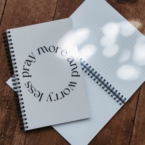 K&ouml;nnte beinhalten: Ein wei&szlig;es Notizbuch mit Spiralbindung und dem Text "worry less pray more" in einem Kreis auf dem Einband.