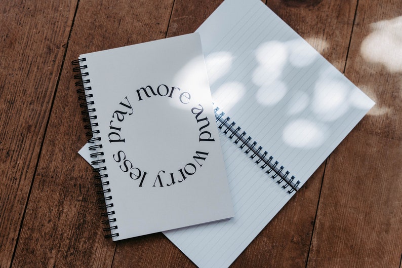 K&ouml;nnte beinhalten: Ein wei&szlig;es Notizbuch mit Spiralbindung und dem Text "worry less pray more" in einem Kreis auf dem Einband.
