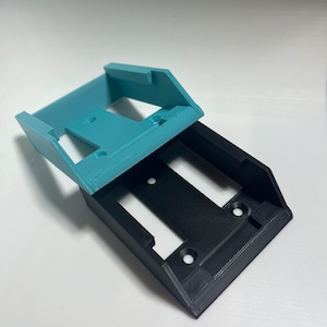 Può includere: Due supporti da parete stampati in 3D, uno nero e uno blu chiaro. I supporti hanno una forma rettangolare con un bordo rialzato in alto e in basso. I supporti hanno diversi piccoli fori sul fondo per il fissaggio a una parete.