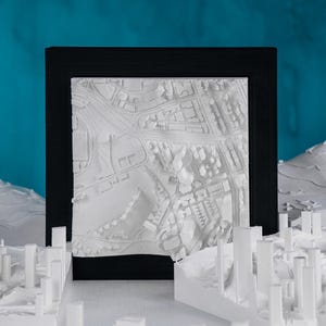 May include: A white 3D printed model of a city skyline in a black frame. The model is detailed and includes buildings, streets, and other features. The model is on a white surface.