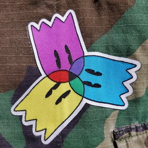 Può includere: Una toppa ricamata colorata con un design geometrico. La toppa è composta da quattro forme sovrapposte: una forma viola, una blu, una gialla e una verde. Ogni forma ha un contorno nero e un bordo bianco.