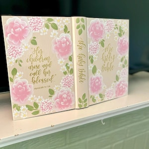 May include: A beige Bible with a hand-painted floral design featuring pink roses, white daisies, and green leaves. The front cover displays the text "Her children arise and call her blessed" and "Proverb 31:28" in gold script. The spine reads "The Holy Bible".