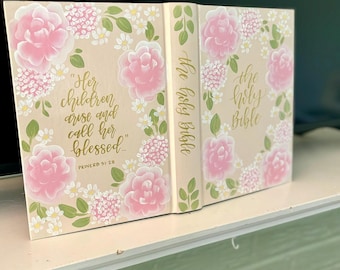 May include: A beige Bible with a hand-painted floral design featuring pink roses, white daisies, and green leaves. The front cover displays the text "Her children arise and call her blessed" and "Proverb 31:28" in gold script. The spine reads "The Holy Bible".