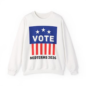 Può includere: Felpa bianca con un design grafico. Il design presenta un rettangolo blu con tre stelle bianche e la parola "VOTE" in bianco. Sotto, strisce rosse e bianche e il testo "MIDTERMS 2026".