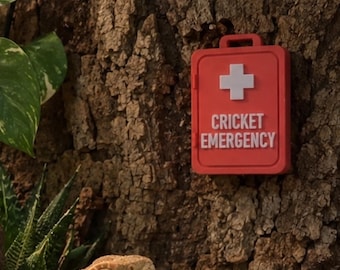 May include: A red, rectangular plastic container mounted on a textured bark wall. The container features a white cross symbol and the white text CRICKET EMERGENCY. A bearded dragon is visible in the lower left corner of the enclosure.