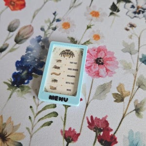 May include: A light blue rectangular frame containing a menu with illustrations of a spider, fruit flies, house crickets, mealworms, bottle flies, and moths. The word MENU is printed in black letters at the bottom of the light blue frame.