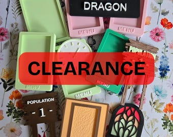 May include: A collection of silicone molds in various shapes and colors, including rectangular menu frames, a clock face, a brown sign reading POPULATION 1, a black sign reading BEWARE OF DRAGON, and a stained glass window design mold.