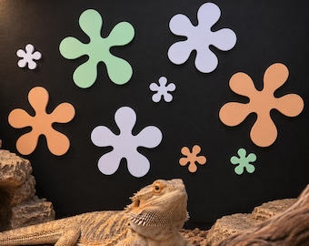 May include: A bearded dragon lizard rests on a sandy substrate in front of a black wall decorated with various sized, flat, cutout flower shapes in shades of light green, white, and peach, arranged in a scattered, non-uniform pattern.