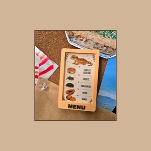 May include: A rectangular light orange sign with the word MENU at the bottom. It features an illustration of a gecko at the top and a list of food items including complete gecko diet, crickets, dubia roaches, papaya, and mango with icons.