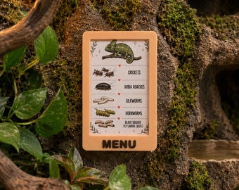 May include: A rectangular beige sign with the word MENU at the bottom. The sign features a green chameleon illustration at the top and a list of feeder insects including crickets, dubia roaches, silkworms, hornworms, and black soldier fly larvae.