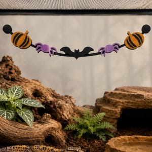 May include: A Halloween-themed hanging decoration featuring a black arched bar with two orange pumpkins on the ends, two purple spiders, and a central black bat. The decoration is suspended horizontally with circular black end mounts.