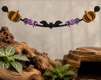 May include: A Halloween-themed hanging decoration featuring a black arched bar with two orange pumpkins on the ends, two purple spiders, and a central black bat. The decoration is suspended horizontally with circular black end mounts.