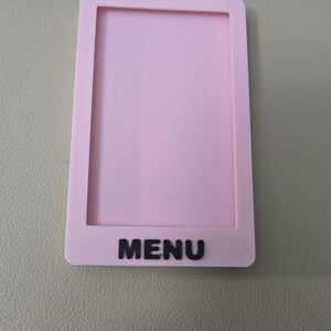 May include: A rectangular light pink plastic frame with a hollow center, featuring the word MENU in raised black capital letters at the bottom. The object has a smooth, matte finish and a simple, clean design with rounded corners on the outer edges.